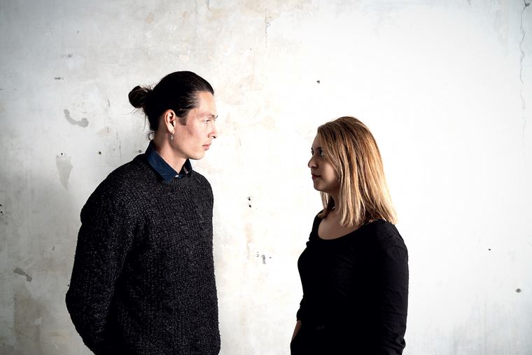 Eda Er and Simon Sieger in profile, facing each other across a plaster wall.